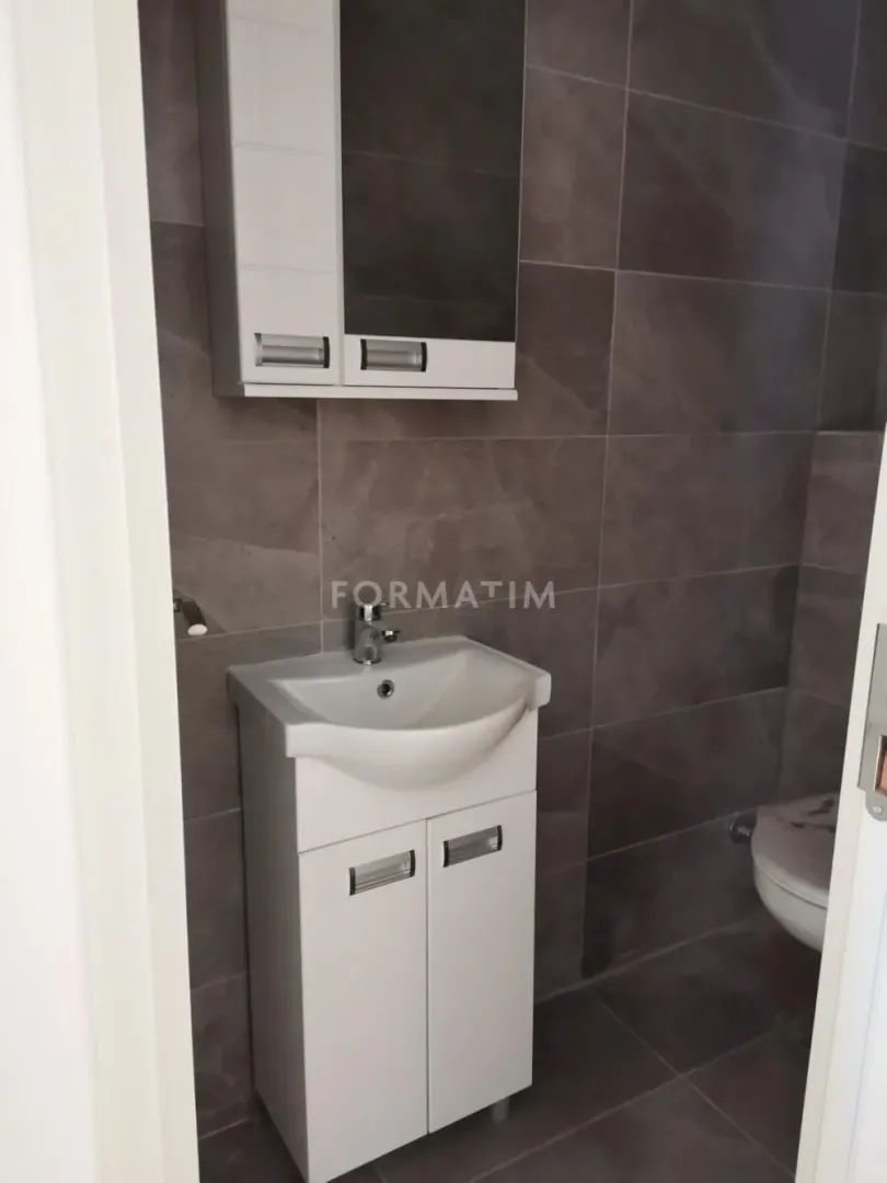 Manje kupatilo sa umivaonikom, ormarićem i toaletom obloženo tamnim pločicama.