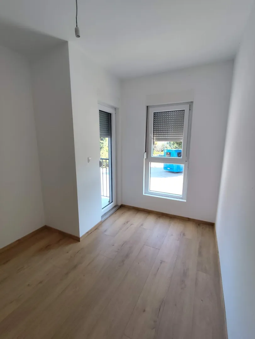 Empty bright room with white walls, wooden flooring, a window, and balcony doors.