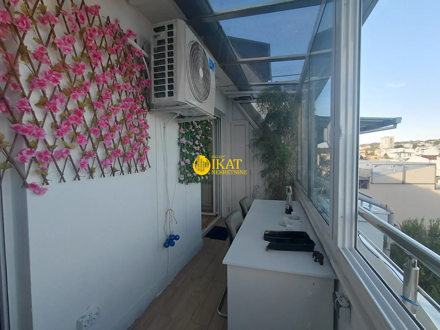 Zastakljeni balkon sa stolom i dekorativnim veštačkim cvećem na zidu.