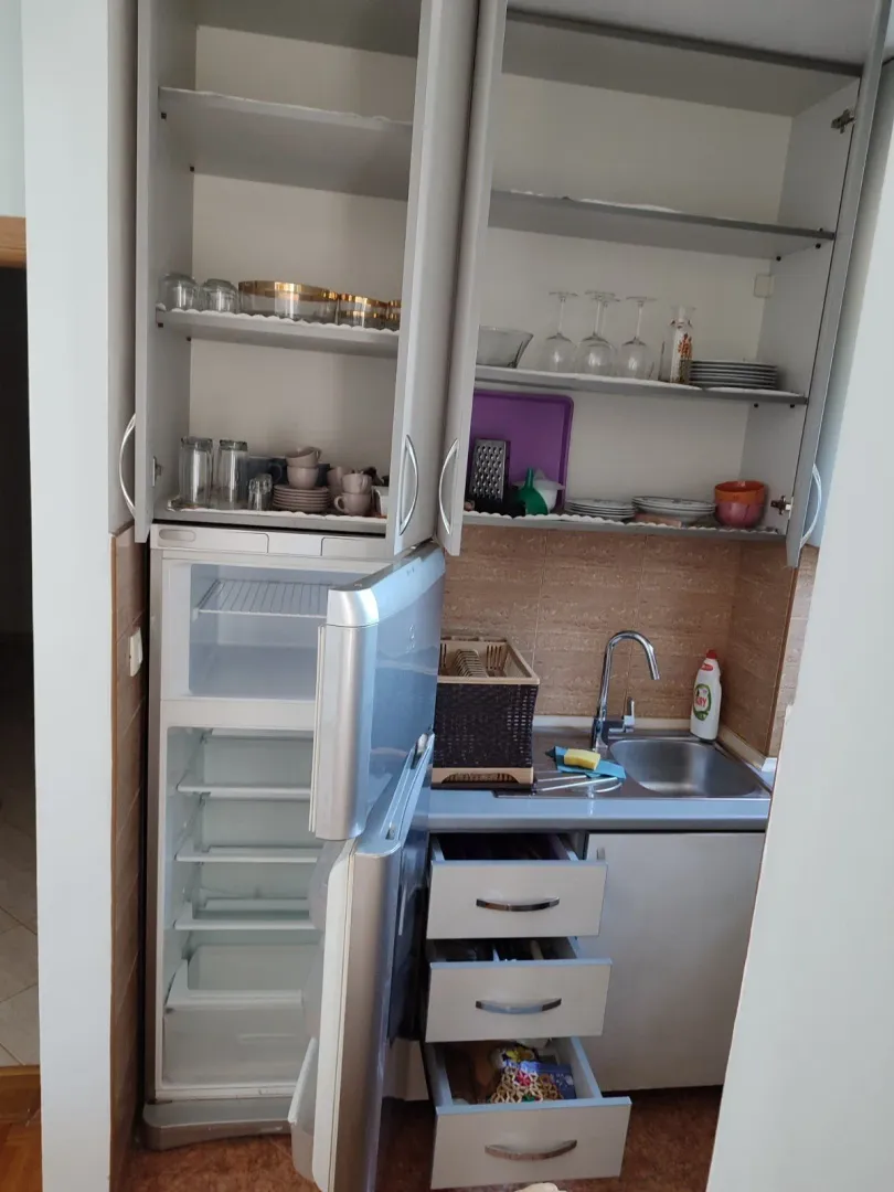 Mala funkcionalna kuhinja sa otvorenim kuhinjskim elementima i frižiderom.