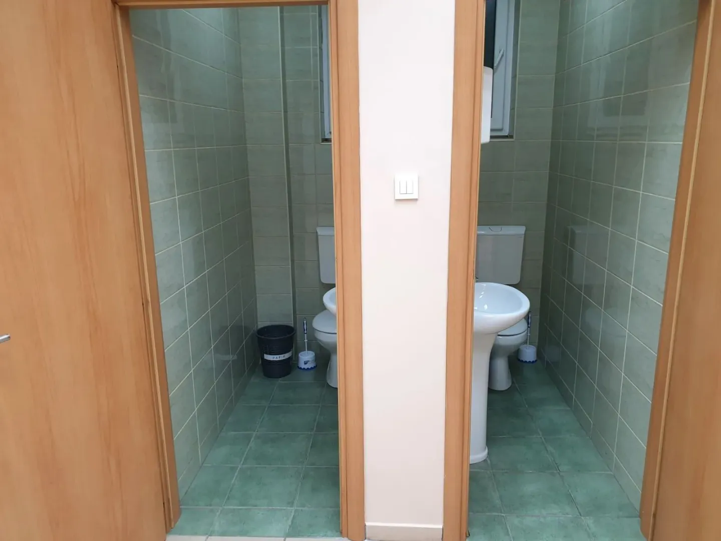 Two toilets with green tiles and wooden doors.