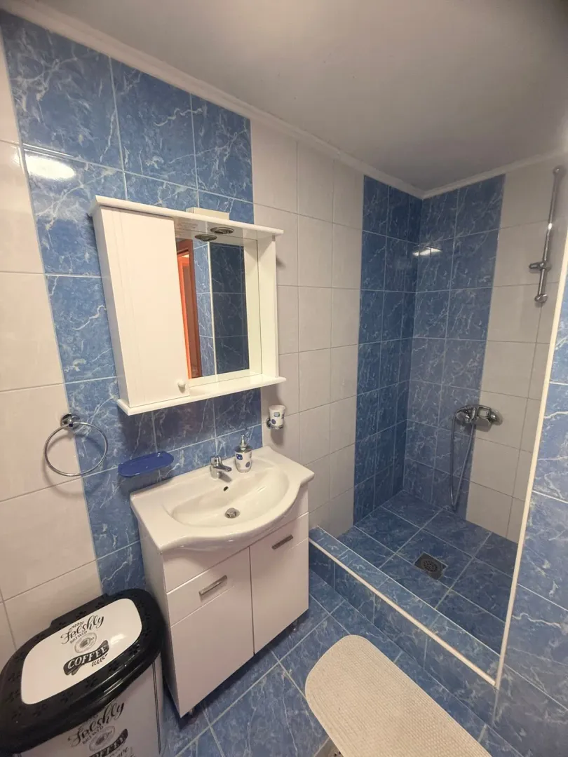 Kupatilo sa umivaonikom, ormarićem sa ogledalom i tuš kabinom obloženo plavim i bijelim pločicama.