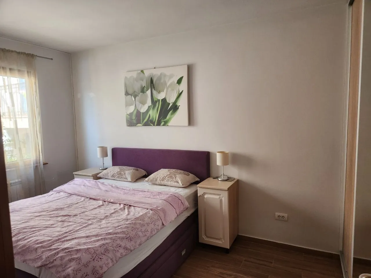 Namještena spavaća soba sa bračnim krevetom, noćnim stočićima i slikom na zidu.