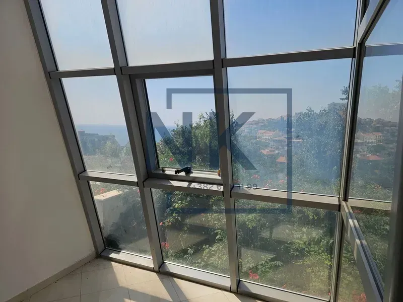 Pogled kroz veliku staklenu stenu na okolno zelenilo i more.