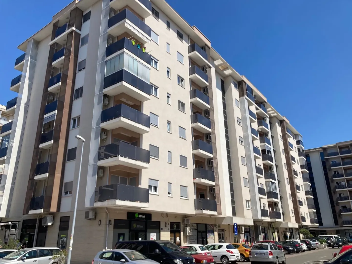 Exterior of a multi-story residential building with balconies and parked cars in front.