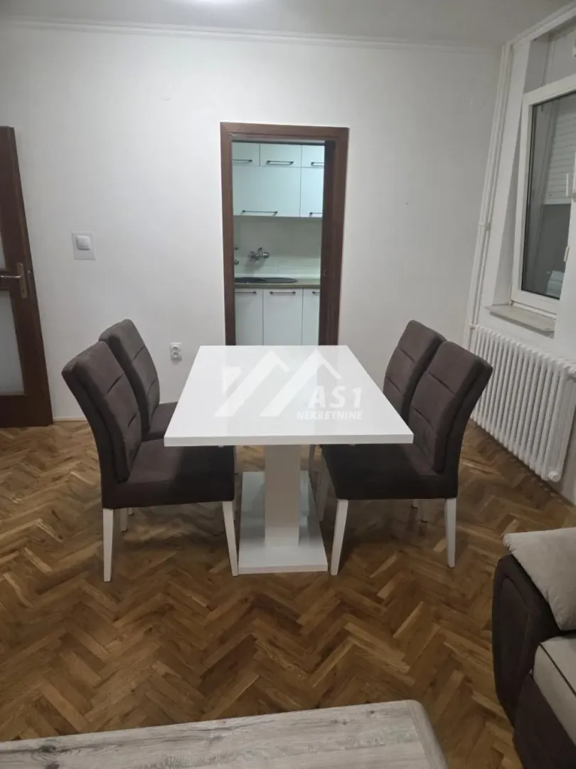 Trpezarija sa belim stolom i četiri tapacirane stolice, sa pogledom na kuhinju.