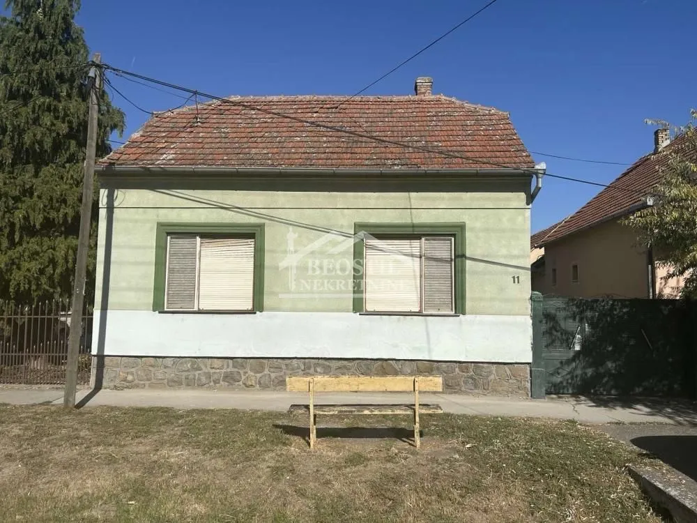 Pogled na prednju fasadu i krov prizemne kuće sa klupom u dvorištu.
