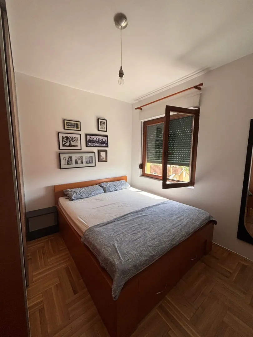 Manja spavaća soba sa bračnim krevetom, drvenim parketom i prozorom.