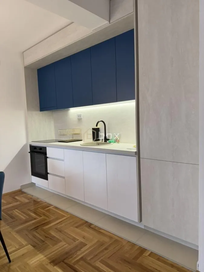 Moderna kuhinja sa belim elementima, plavim gornjim ormarićima i ugradnim uređajima.