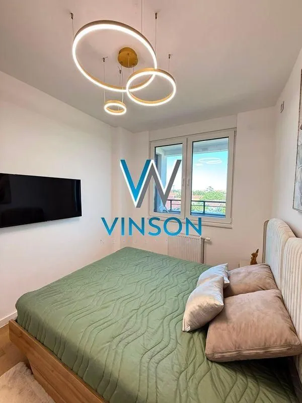 Nameštena spavaća soba sa bračnim krevetom, televizorom i modernim lusterom.