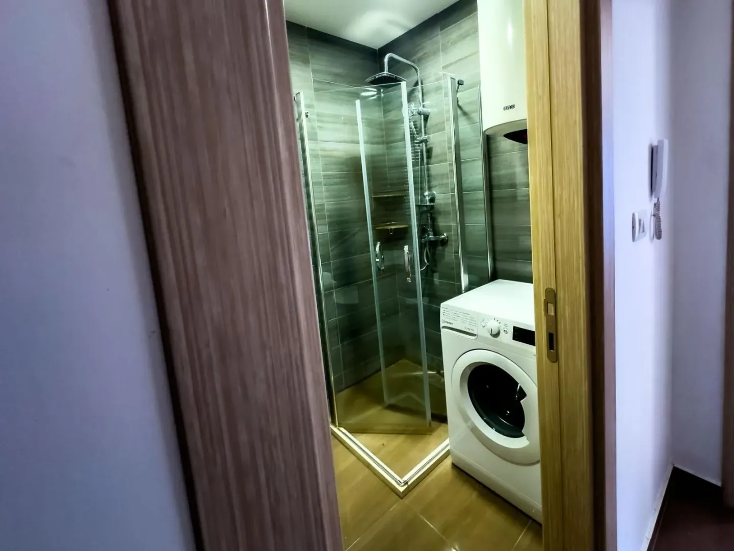 Pogled kroz otvorena vrata na kupatilo sa tuš kabinom i veš mašinom.