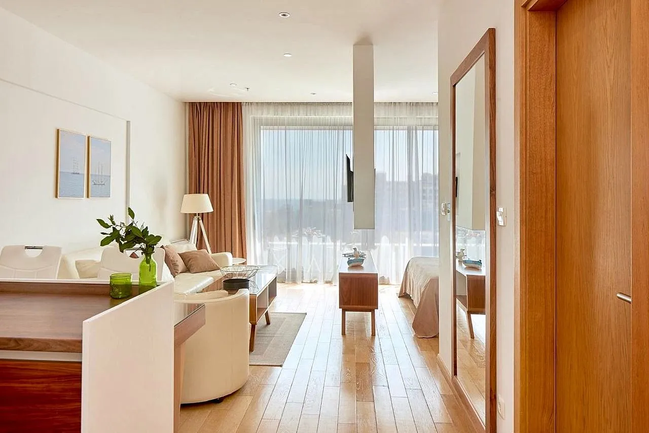 View from the hallway into a bright living room with wooden flooring and large windows.