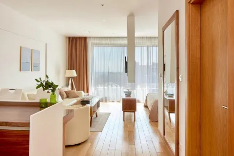 View from the hallway into a bright living room with wooden flooring and large windows. - image 3
