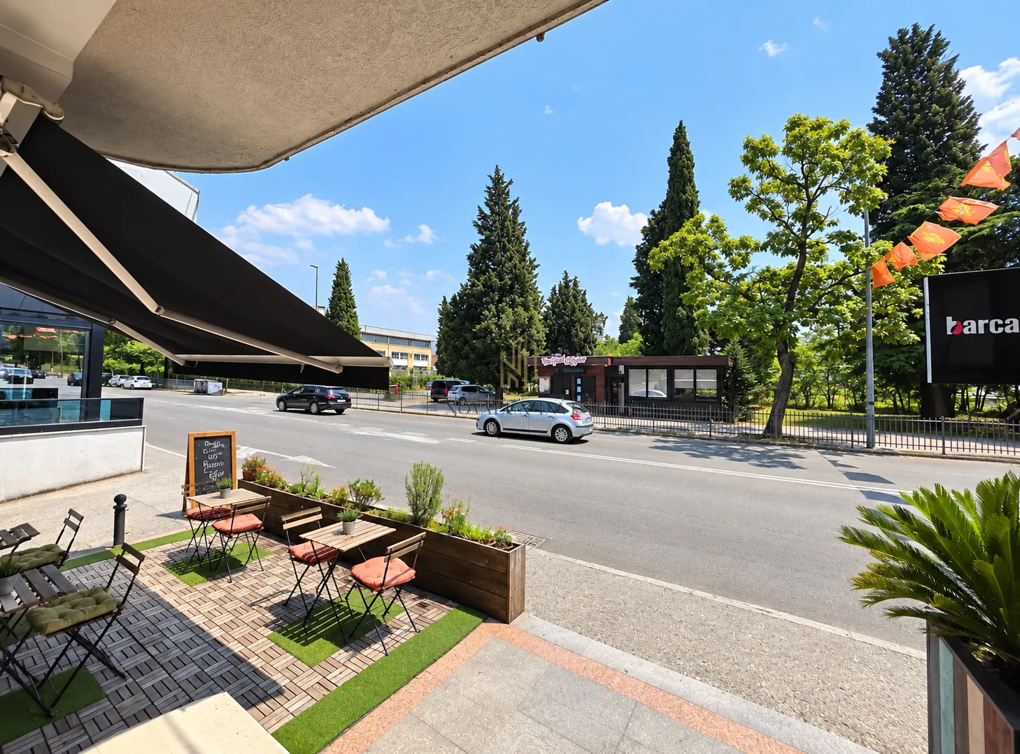 Outdoor cafe terrace with tables and a view of the street and green areas.