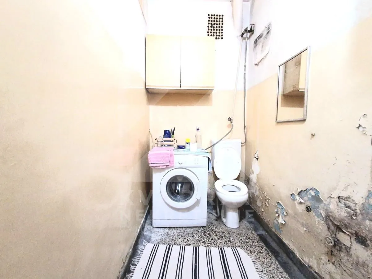 Starije kupatilo sa toaletom, veš mašinom i oštećenim zidovima.