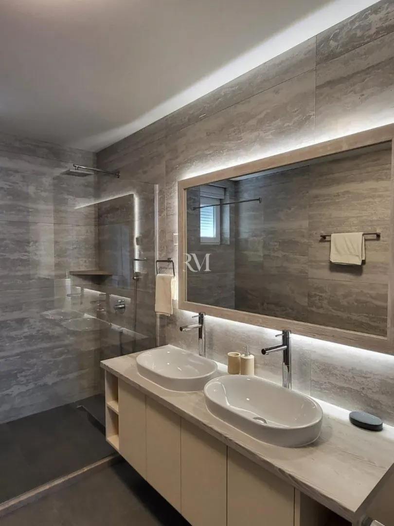 Modern bathroom with double sinks, lighted mirror and walk-in shower.