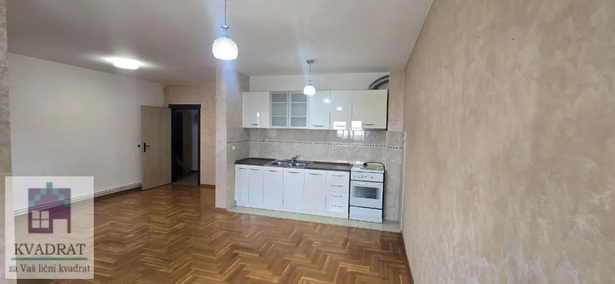 Manja kuhinja sa belim elementima i šporetom u sklopu prostorije sa parketom.