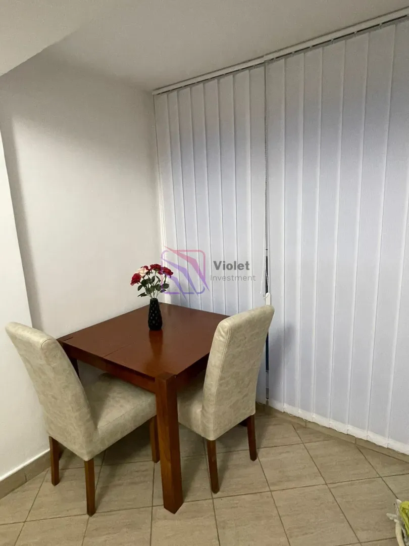 Small wooden dining table with two chairs near a window with vertical blinds.