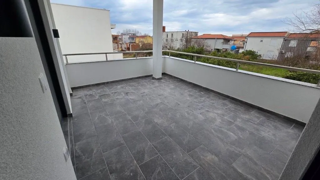 Prostrana popločana terasa sa metalnom ogradom i pogledom na naselje.