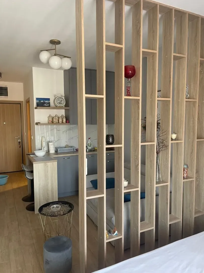 Living room with a wooden partition shelving unit and a kitchen in the background.