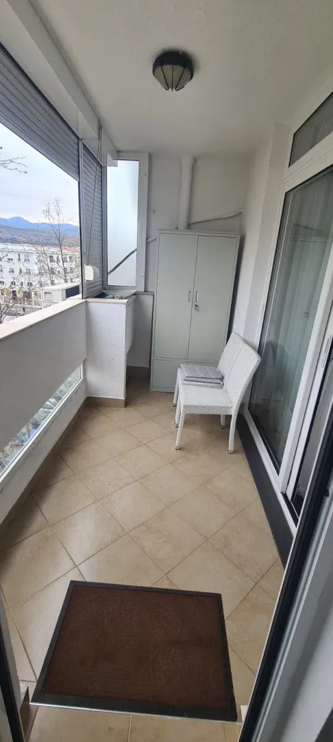 Uski zatvoreni balkon sa keramičkim pločicama, bijelim stolicama i metalnim ormarom.