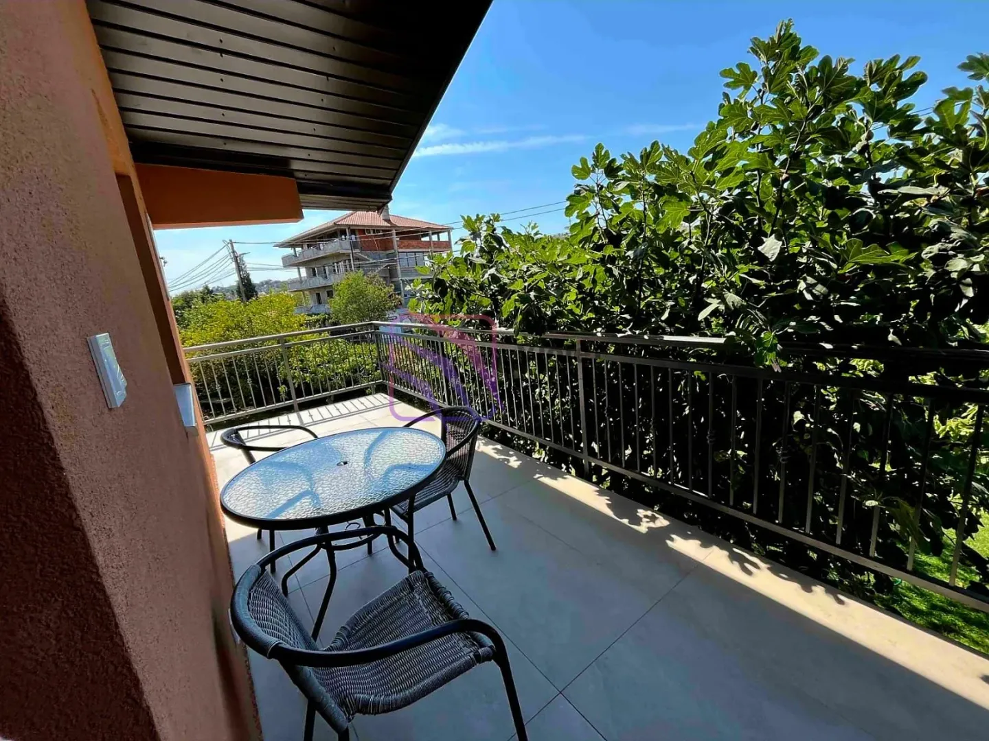 Natkrivena terasa sa stolom i stolicama i pogledom na zelenilo.