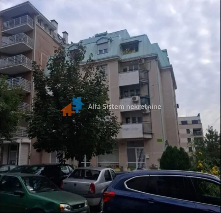 Pogled na spoljašnjost stambene zgrade sa parkingom u prvom planu.