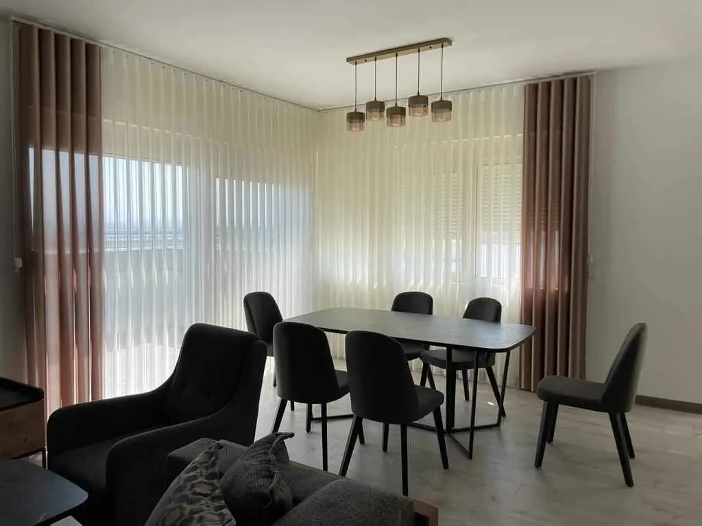 Svijetla trpezarija sa modernim stolom, stolicama i elegantnim zavjesama.