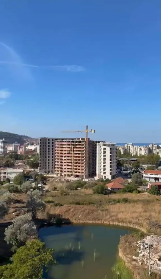 View of a building under construction and the surrounding landscape with a small water basin in the foreground.