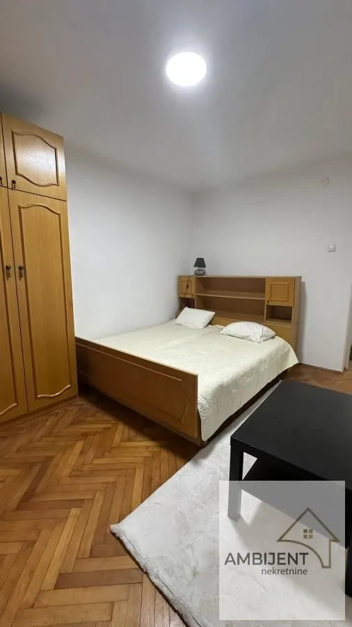 Nameštena spavaća soba sa bračnim krevetom, drvenim ormarom i parketom.