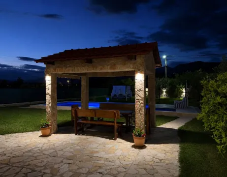 Illuminated stone gazebo in the yard with a dining table and a swimming pool in the background. - image 3