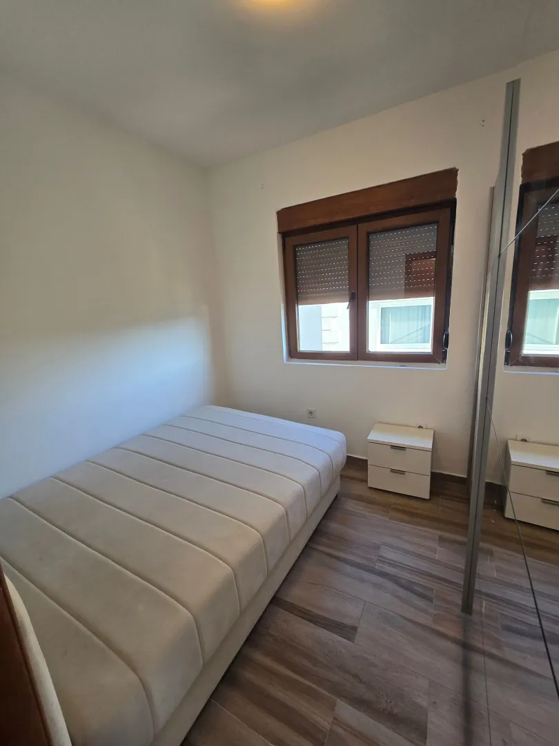 Manja spavaća soba sa krevetom, noćnim stočićem i prozorom.