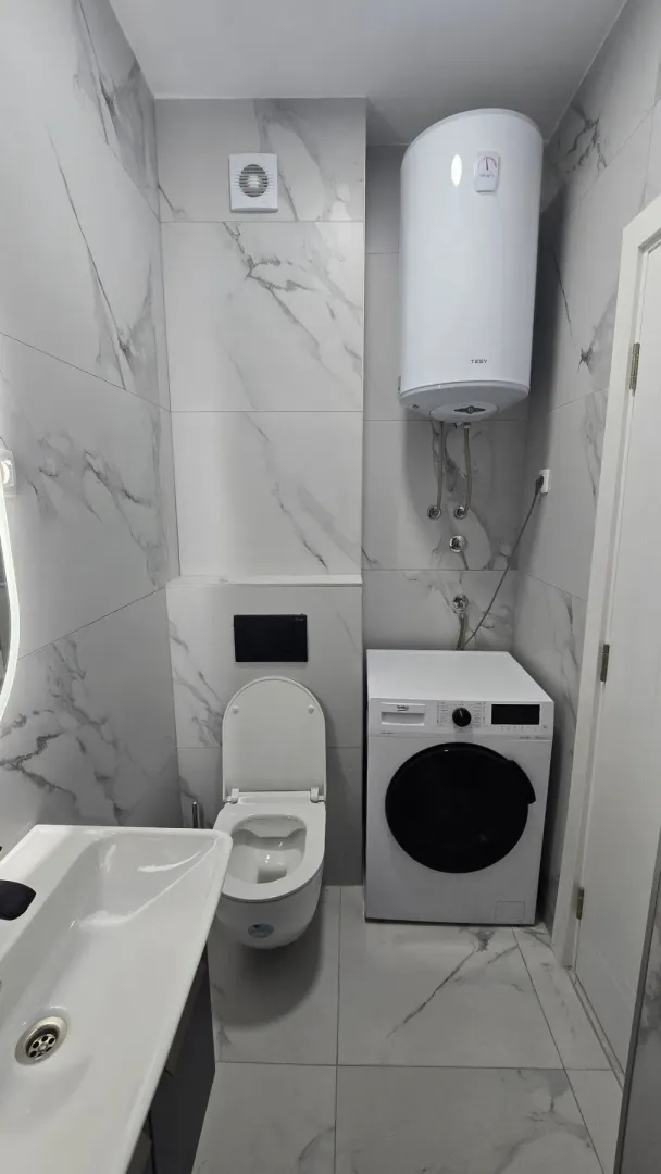 Moderno opremljeno kupatilo sa bijelim mermernim pločicama, wc šoljom, umivaonikom i veš mašinom.