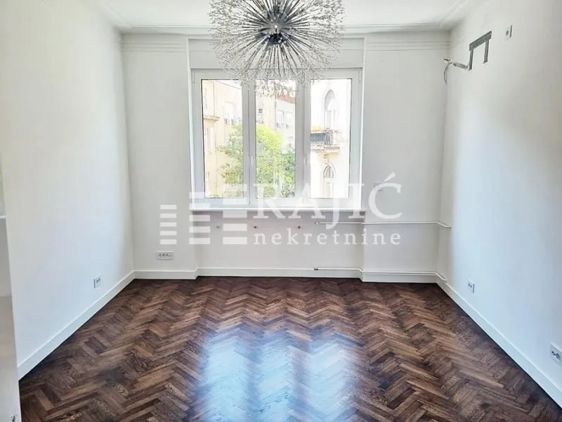 Empty room with hardwood parquet flooring and a large window.