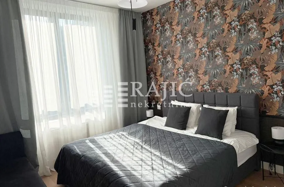 Furnished bedroom with a double bed, patterned wallpaper, and a window with curtains.