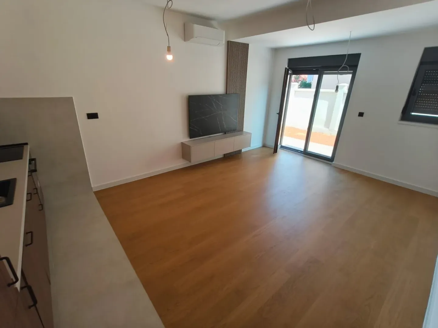 Prazan dnevni boravak sa drvenim podom, TV komodom i izlazom na terasu.