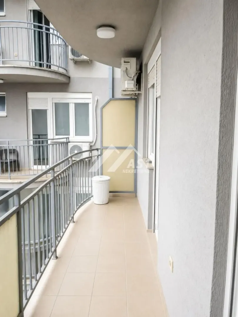 Pogled na duguljasti popločani balkon sa metalnom ogradom i spoljnom jedinicom klima uređaja.