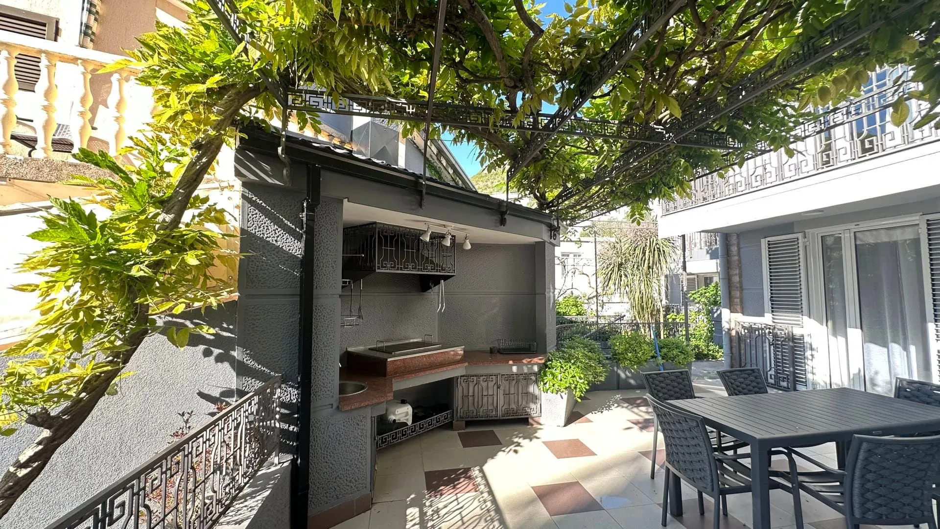 A paved patio featuring an outdoor kitchen, barbecue, and dining set under a vine-covered pergola.