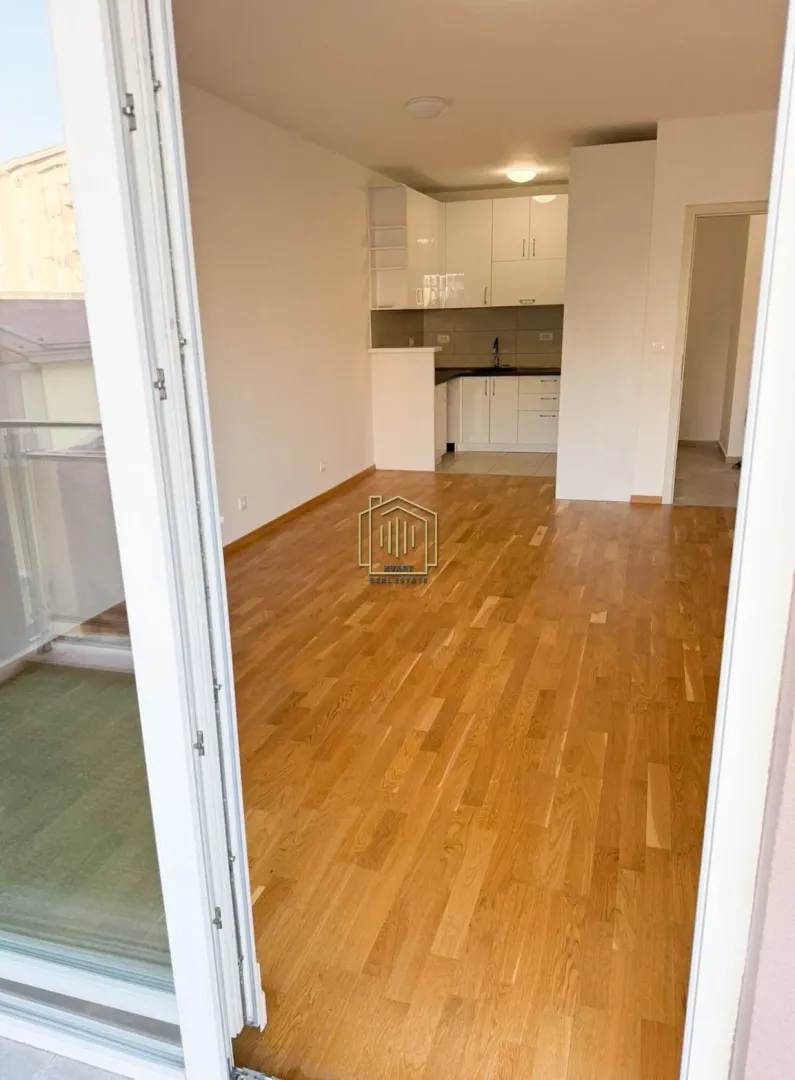 Empty living room with wooden flooring and a view of the kitchen.