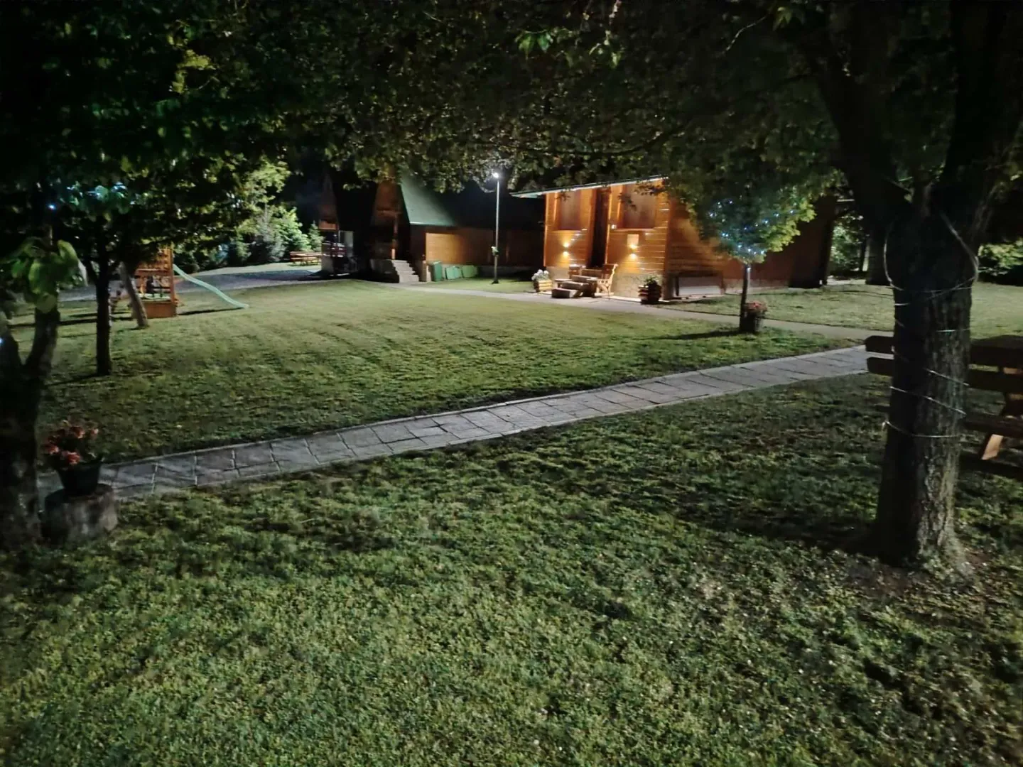 Noćni pogled na osvetljeno dvorište i drvenu kuću sa stazom.