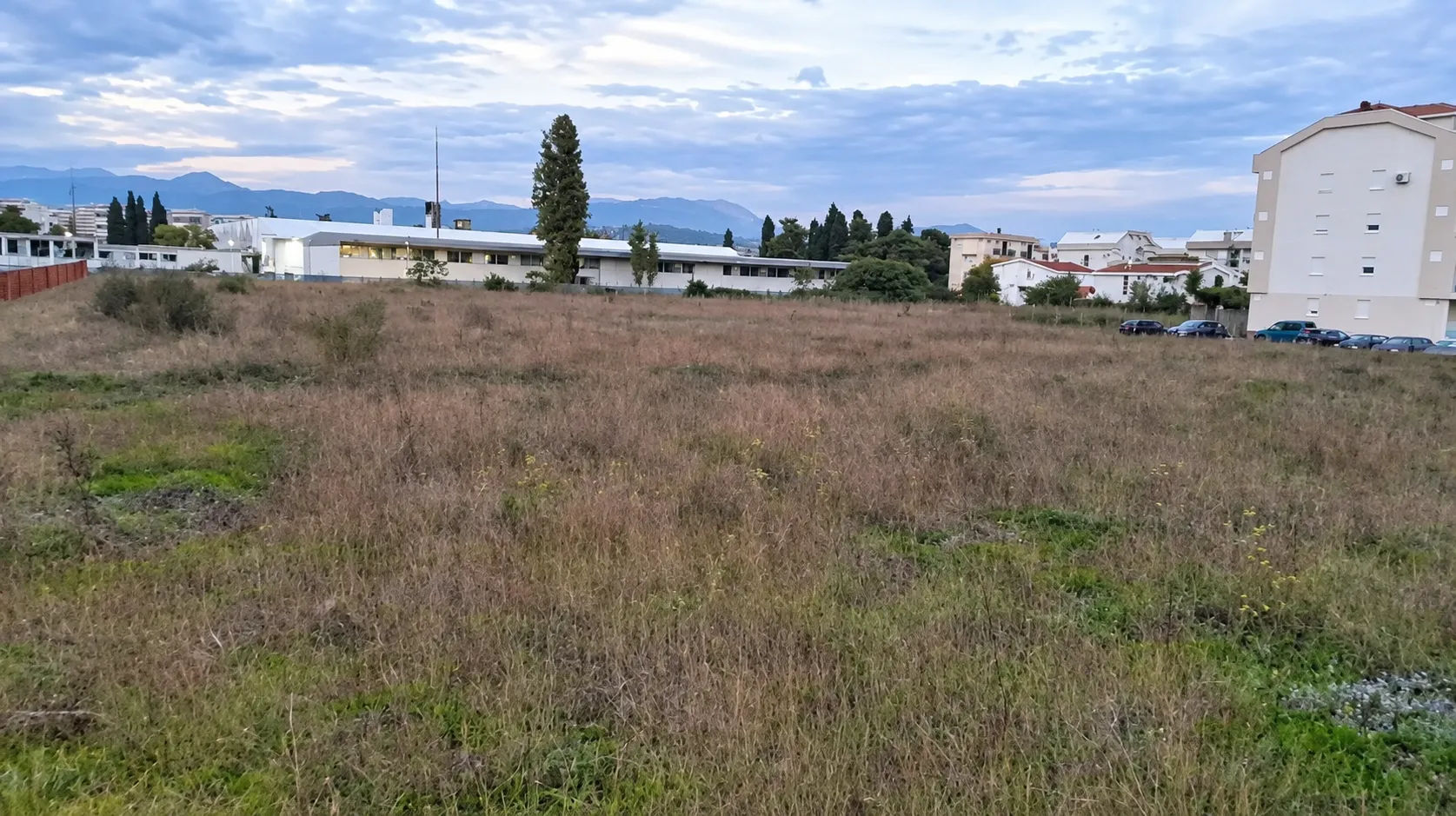 Pogled na prostrano neizgrađeno zemljište sa okolnim zgradama u daljini.