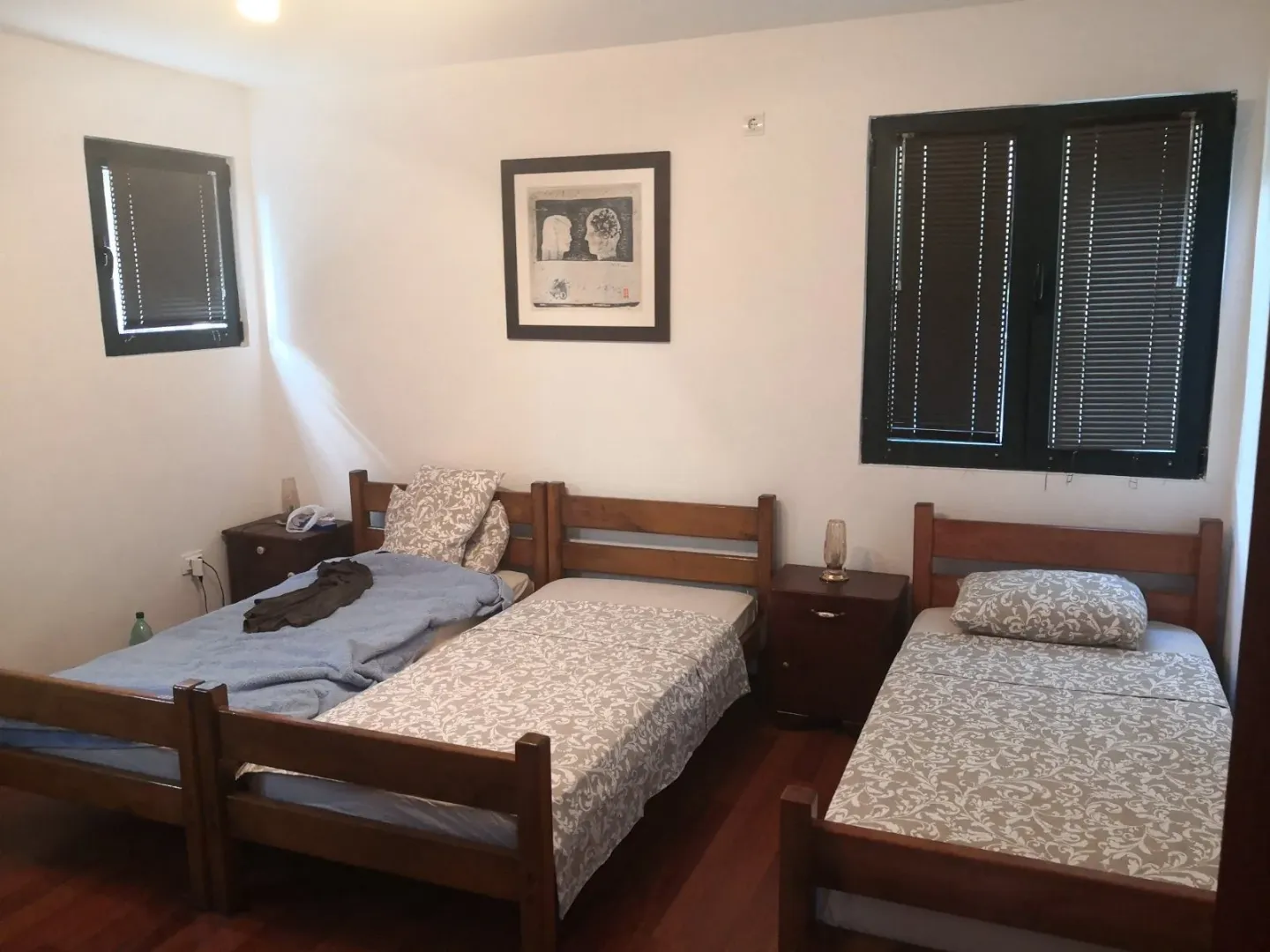 Spavaća soba sa tri drvena kreveta, noćnim ormarićima i prozorima sa roletnama.