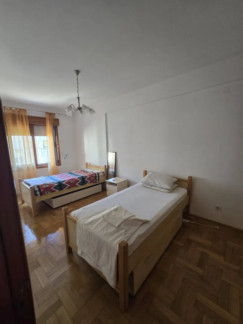 Spavaća soba sa dva drvena kreveta, parketom i prozorom sa žutim zavesama.