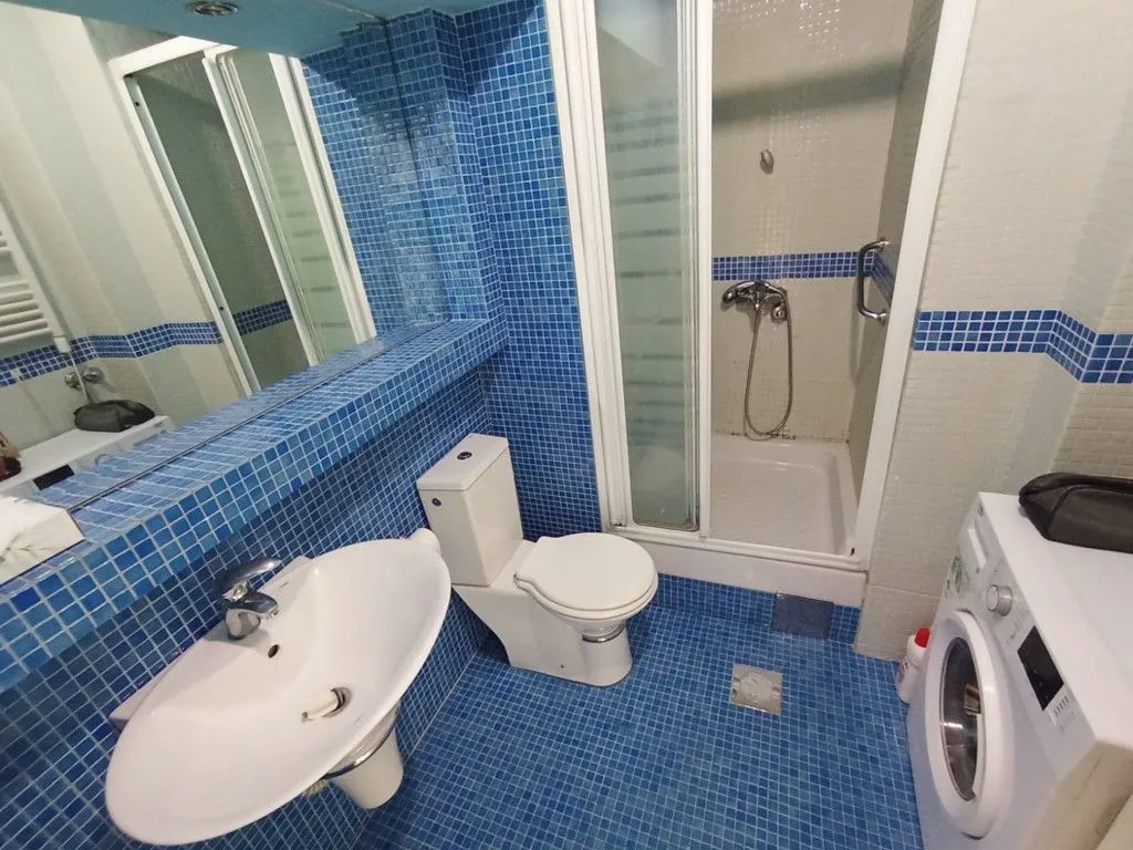 Kupatilo sa plavim mozaik pločicama, tuš kabinom, umivaonikom i veš mašinom.