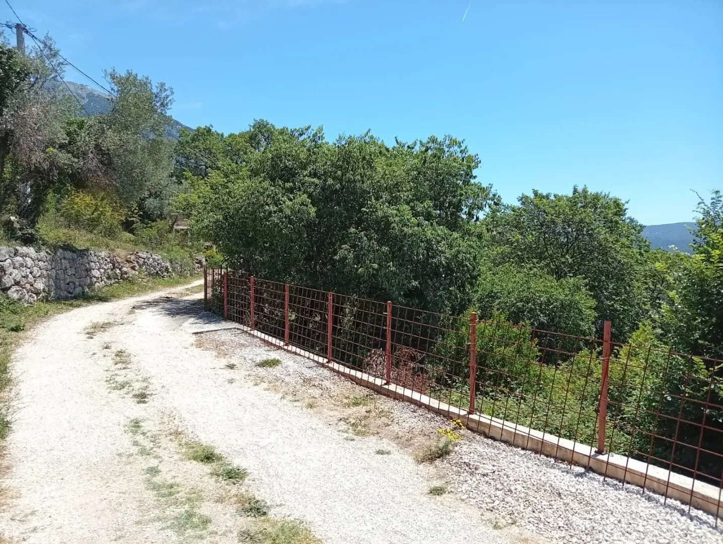 Zemljani put pored impe ograđene metalnom ogradom sa zelenilom.