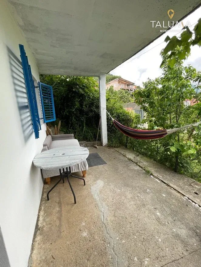 Natkrivena terasa sa stolom, sofom i ležaljkom okružena zelenilom.