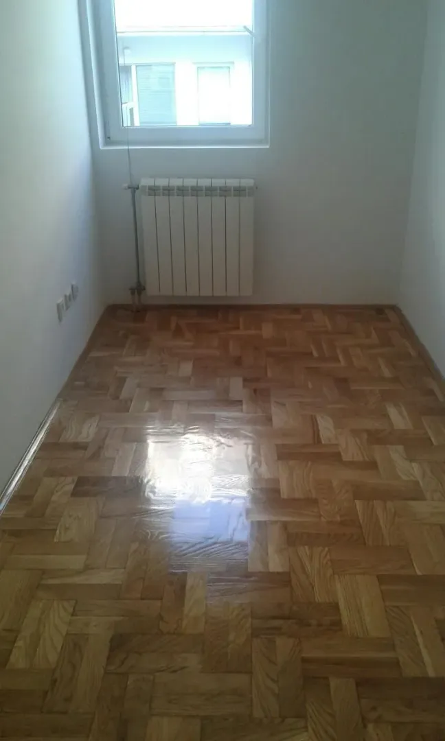 Prazna soba sa parketom i radijatorom ispod prozora.