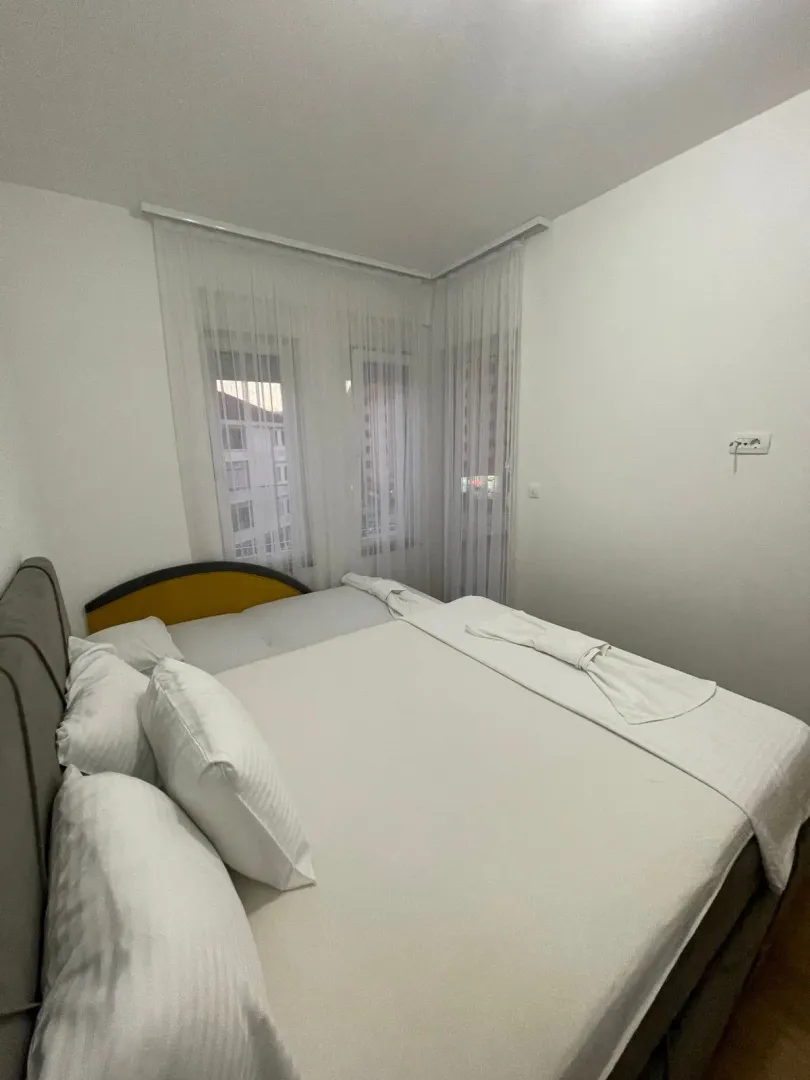 Spavaća soba sa bračnim krevetom i prozorima prekrivenim bijelim zavjesama.