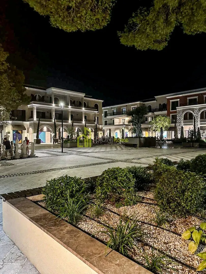 Noćni pogled na osvijetljeni trg sa okolnim zgradama i uređenim rastinjem u kompleksu.