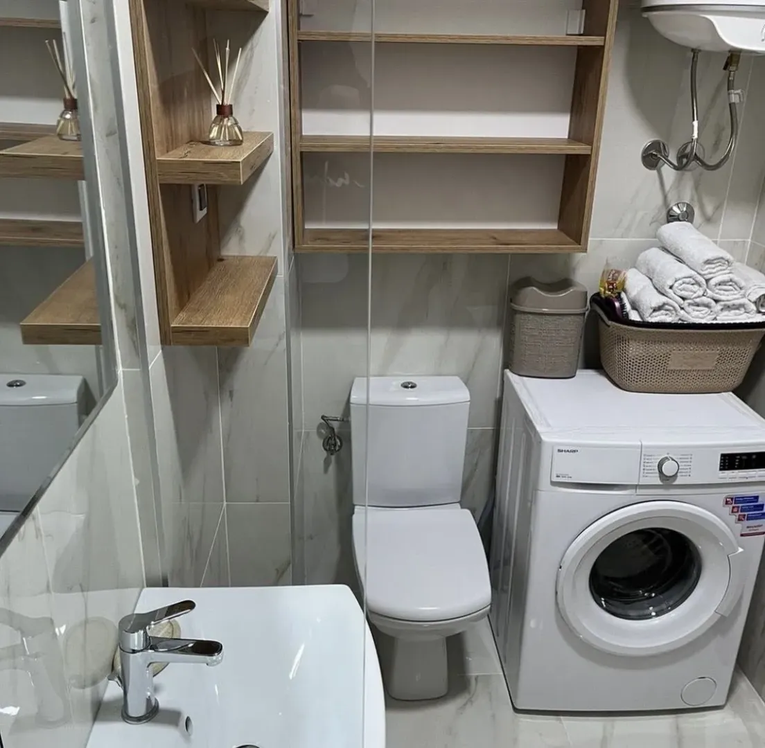 Moderno kupatilo sa veš mašinom, toaletom i drvenim policama.